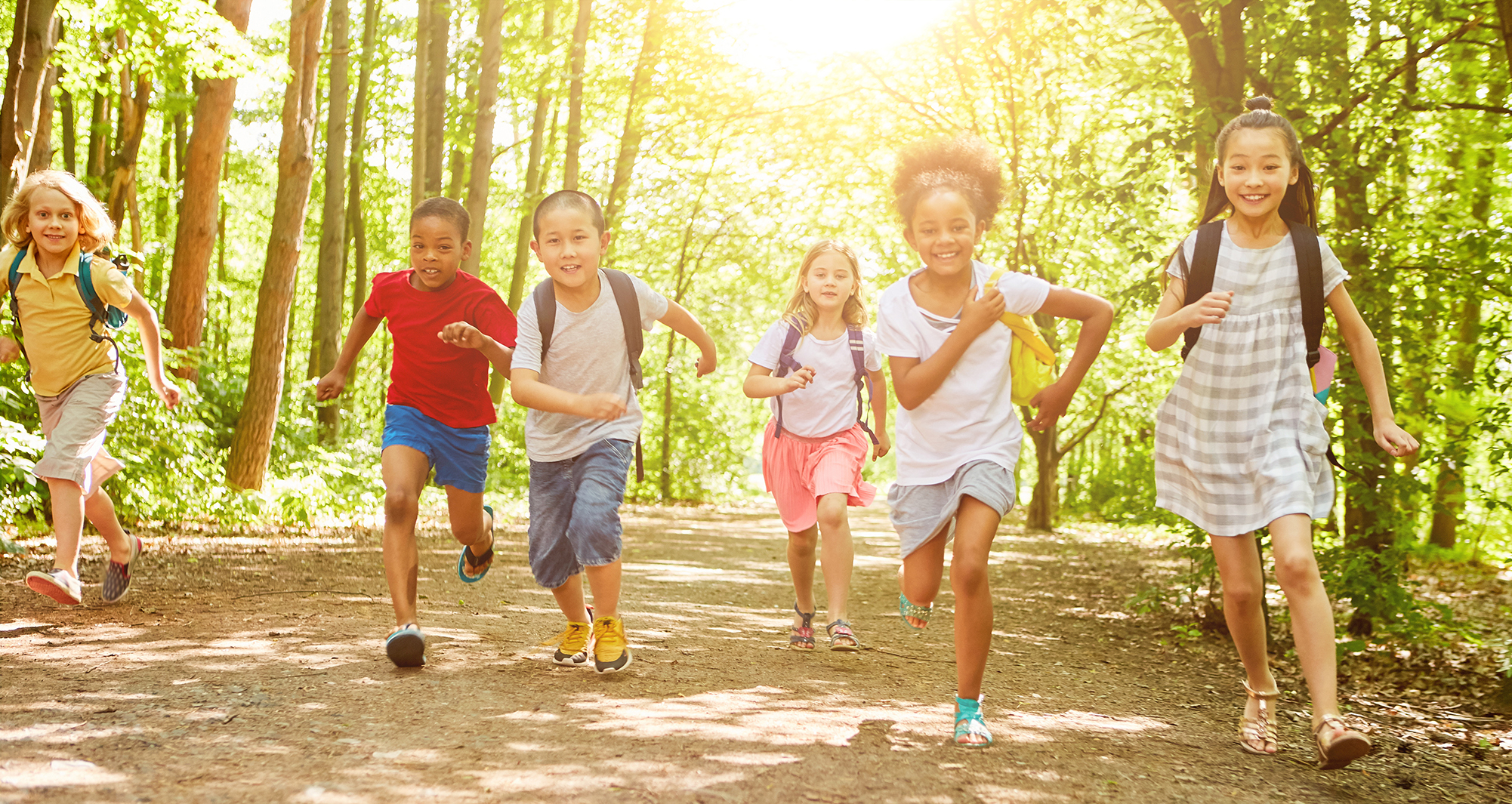 Children running down a trail through the forest. They are smiling and the sun is shining behind them.
