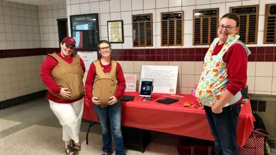Three staff members wear fake pregnancy bellies in front of their fair booth.